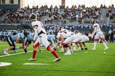 High school football game in progress with players in light blue and red uniforms competing on field during daytime match.