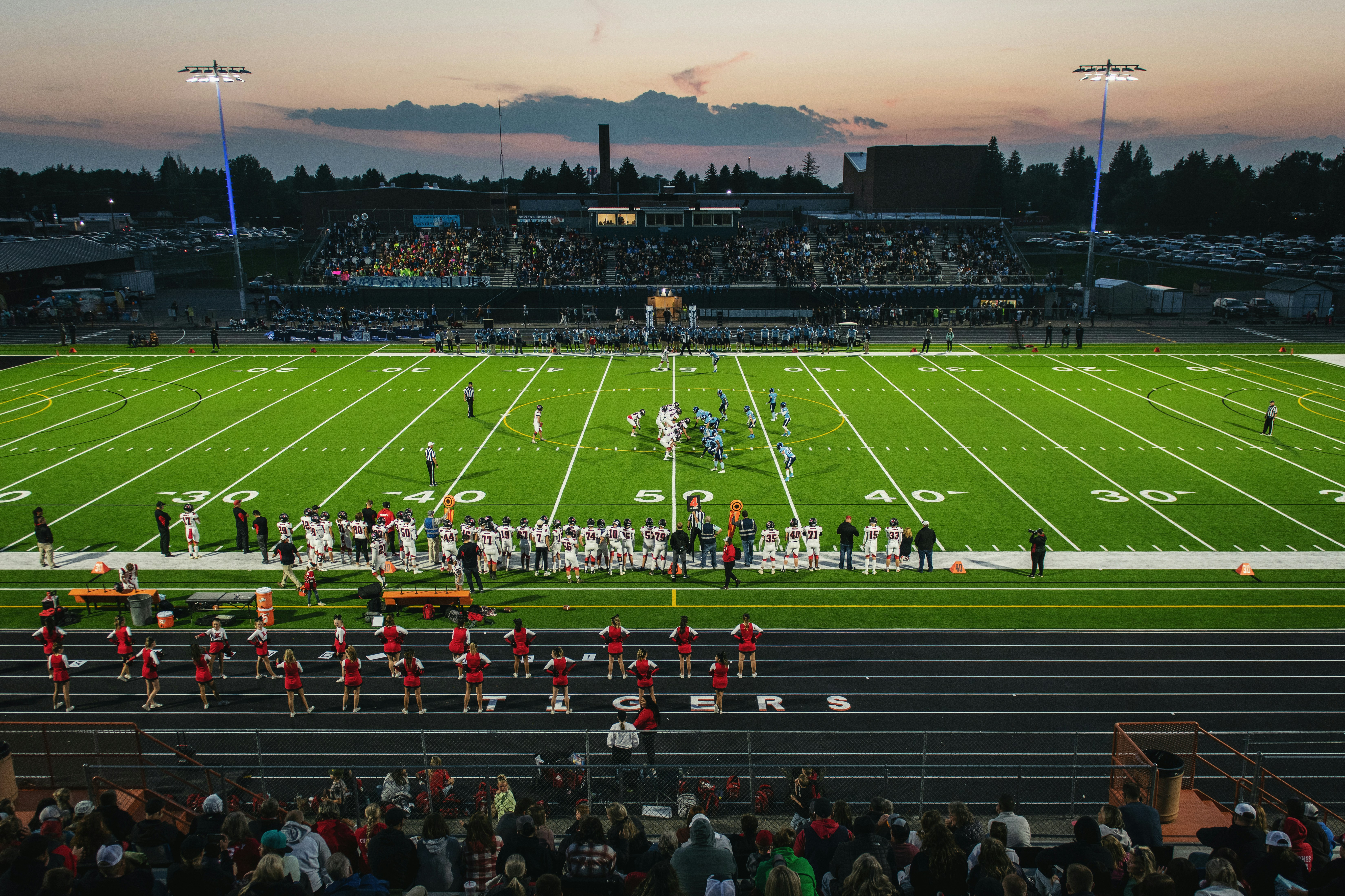 Evening football game at stadium with illuminated field, marching band on track, and crowd in stands at dusk