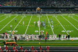 Evening football game at stadium with illuminated field, marching band on track, and crowd in stands at dusk