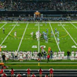 Evening football game at stadium with illuminated field, marching band on track, and crowd in stands at dusk