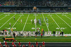 Evening football game at stadium with illuminated field, marching band on track, and crowd in stands at dusk