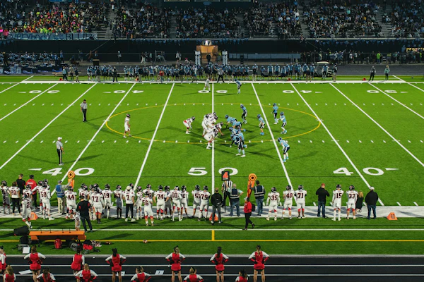 Evening football game at stadium with illuminated field, marching band on track, and crowd in stands at dusk