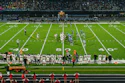 Evening football game at stadium with illuminated field, marching band on track, and crowd in stands at dusk